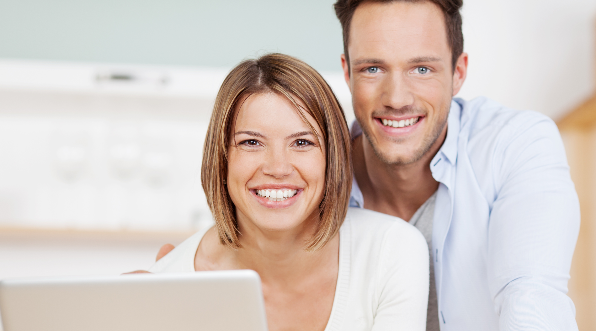 Eine Frau und ein Mann sitzen beieinander und blicken über einen Laptop hinweg Richtung Kamera 